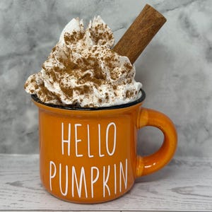 May include: Orange mug with the text "HELLO PUMPKIN" in white. The mug is filled with whipped cream, sprinkled with cinnamon, and topped with a cinnamon stick. The mug has a black rim and a curved handle.