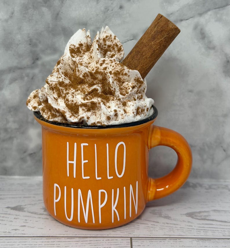 May include: Orange mug with the text "HELLO PUMPKIN" in white. The mug is filled with whipped cream, sprinkled with cinnamon, and topped with a cinnamon stick. The mug has a black rim and a curved handle.