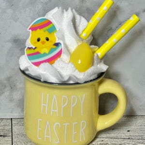 Op de afbeelding: Een gele keramische mok met de tekst "Happy Easter" aan de voorkant. De mok is gevuld met witte slagroom, een geel snoepje ei en een kleurrijk kuiken in een ei. Twee geel-wit gestreepte rietjes steken uit de slagroom.