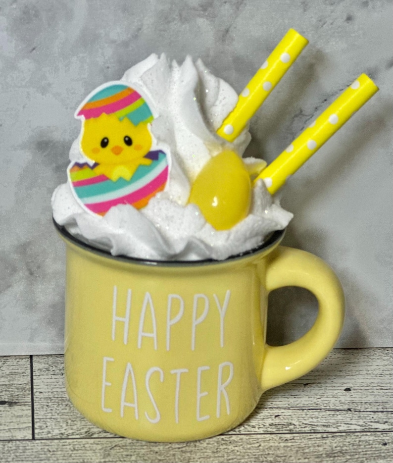 Op de afbeelding: Een gele keramische mok met de tekst "Happy Easter" aan de voorkant. De mok is gevuld met witte slagroom, een geel snoepje ei en een kleurrijk kuiken in een ei. Twee geel-wit gestreepte rietjes steken uit de slagroom.