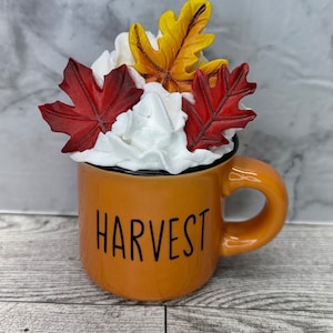May include: An orange mug with the word "HARVEST" in black lettering, filled with white whipped cream and topped with red and yellow fall leaves. The mug has a black rim and a curved handle.