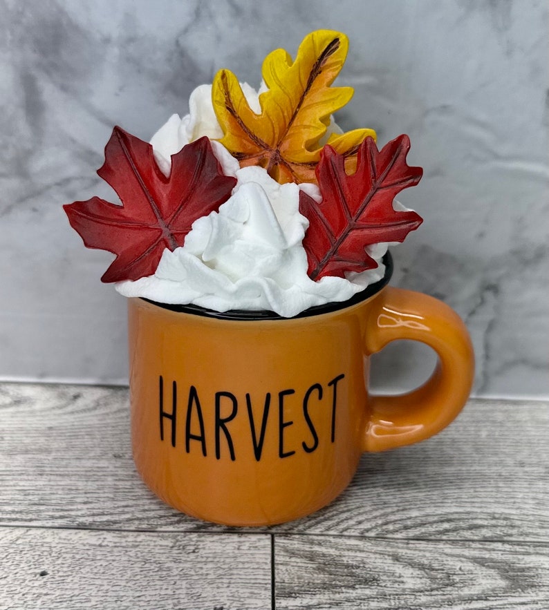 May include: An orange mug with the word "HARVEST" in black lettering, filled with white whipped cream and topped with red and yellow fall leaves. The mug has a black rim and a curved handle.