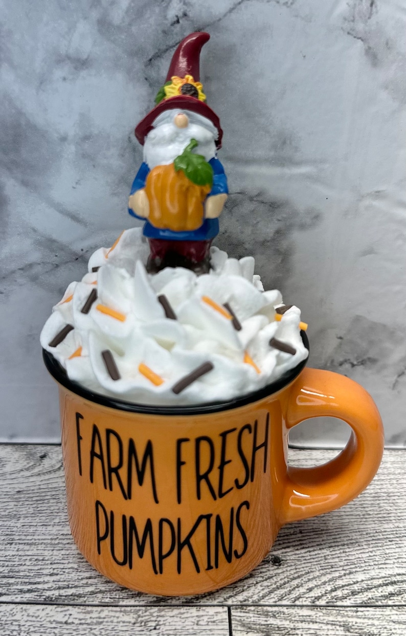 May include: An orange mug with the text "FARM FRESH PUMPKINS" features a gnome figurine holding a pumpkin, atop a whipped cream design with sprinkles. The gnome wears a red hat and blue jacket. A fall-themed decorative item.