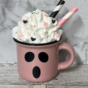 May include: A pink ceramic mug with a ghost face design, filled with whipped cream and topped with pink and black sprinkles. Two striped straws stick out of the whipped cream.