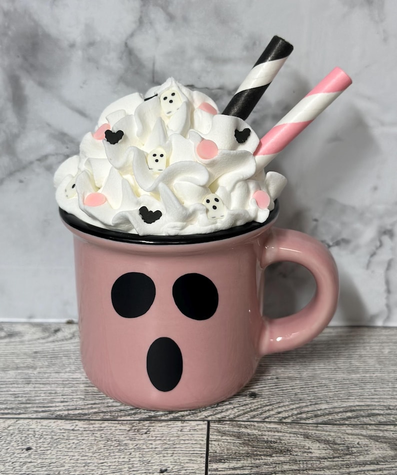 May include: A pink ceramic mug with a ghost face design, filled with whipped cream and topped with pink and black sprinkles. Two striped straws stick out of the whipped cream.