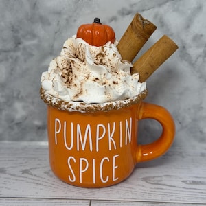 May include: Orange ceramic mug with the words "PUMPKIN SPICE" in white. The mug is topped with whipped cream, a small pumpkin, cinnamon sticks, and a dusting of brown spice. The mug has a handle and is set on a light wood surface.