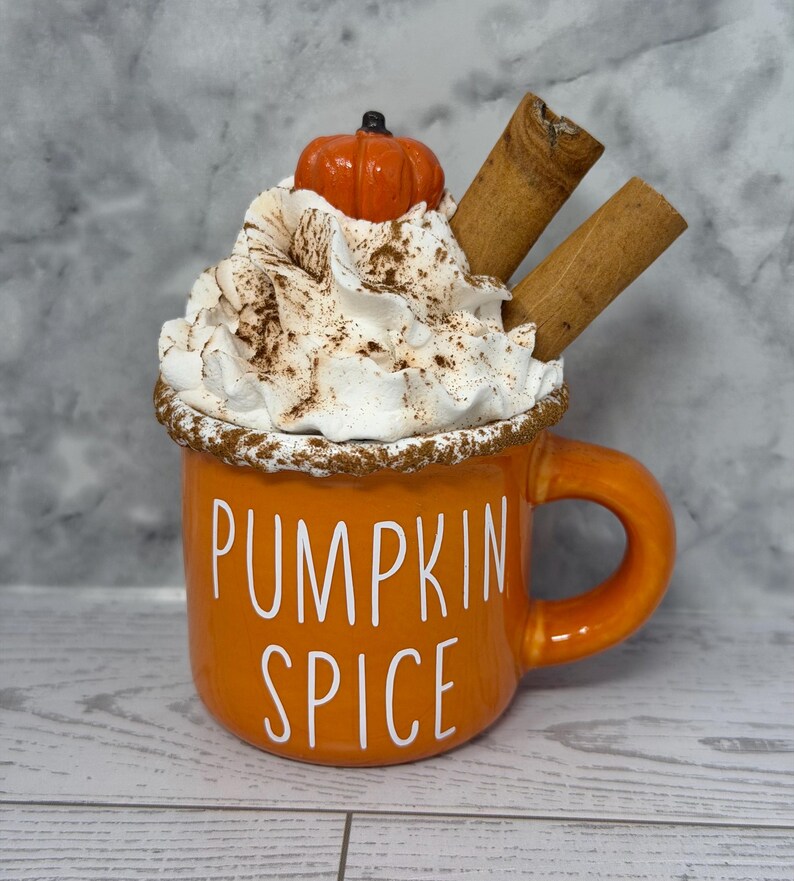 May include: Orange ceramic mug with the words "PUMPKIN SPICE" in white. The mug is topped with whipped cream, a small pumpkin, cinnamon sticks, and a dusting of brown spice. The mug has a handle and is set on a light wood surface.