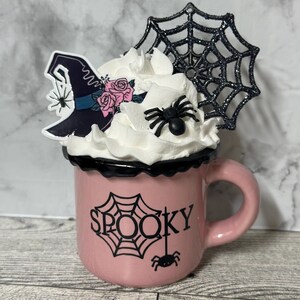 May include: Pink ceramic mug with a black rim and a black spider web design. The mug has the word "SPOOKY" written on it. The mug is topped with whipped cream, a black spider web, a black spider, and a witch's hat with pink flowers.