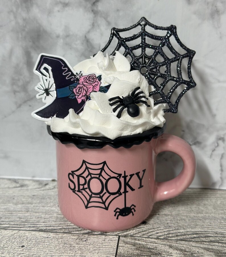 May include: Pink ceramic mug with a black rim and a black spider web design. The mug has the word "SPOOKY" written on it. The mug is topped with whipped cream, a black spider web, a black spider, and a witch's hat with pink flowers.