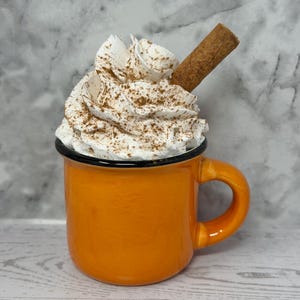 May include: An orange enamel mug filled with whipped cream, dusted with cinnamon, and topped with a cinnamon stick. The mug has a black rim and handle. The background is a marbled gray and white.