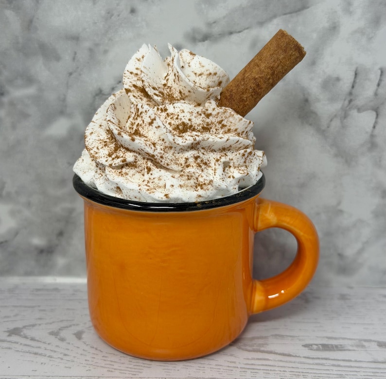 May include: An orange enamel mug filled with whipped cream, dusted with cinnamon, and topped with a cinnamon stick. The mug has a black rim and handle. The background is a marbled gray and white.