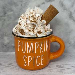 May include: An orange ceramic mug with the words "PUMPKIN SPICE" in white lettering. The mug is filled with whipped cream, sprinkled with cinnamon, and topped with a cinnamon stick. The mug has a black rim and a handle.