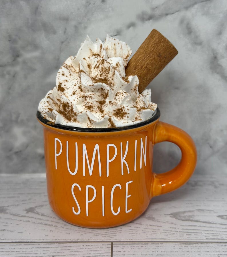 May include: An orange ceramic mug with the words "PUMPKIN SPICE" in white lettering. The mug is filled with whipped cream, sprinkled with cinnamon, and topped with a cinnamon stick. The mug has a black rim and a handle.