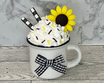 May include: White ceramic mug with a black rim and a black and white gingham bow. The mug is filled with white whipped cream topped with yellow and brown sprinkles and a yellow sunflower.