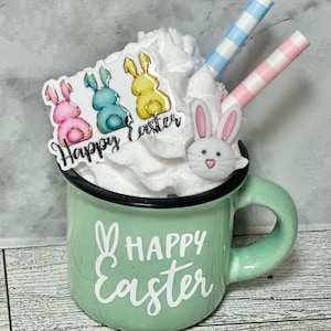 Op de afbeelding: Een groene keramische mok met een witte handgreep en de tekst "Happy Easter" aan de voorkant. De mok is gevuld met slagroom en bedekt met drie konijnenvormige snoepjes en twee gestreepte rietjes.