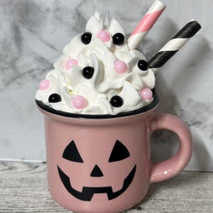 May include: A pink ceramic mug with a jack-o'-lantern face. The mug is filled with whipped cream and topped with pink and black sprinkles. Two striped straws stick out of the whipped cream.
