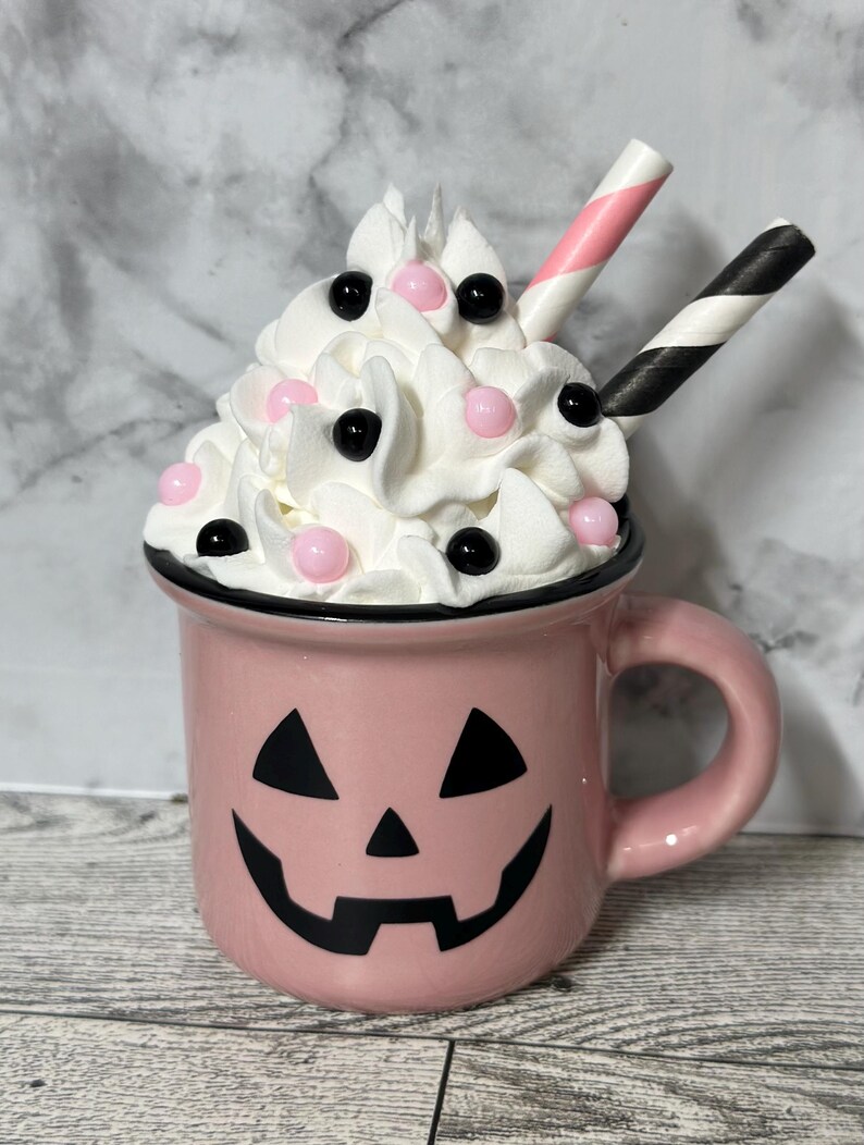 May include: A pink ceramic mug with a jack-o'-lantern face. The mug is filled with whipped cream and topped with pink and black sprinkles. Two striped straws stick out of the whipped cream.