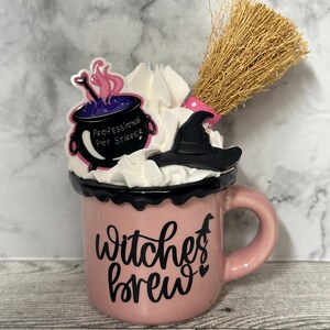 May include: Pink ceramic mug with a black rim and handle. The mug has a black and white graphic that says "Witches Brew" with a small witch silhouette and two hearts. The mug is topped with whipped cream, a black witch's hat, and a small purple cauldron with a pink and white "Professional Pot Stirrer" sticker. A brown broom is sticking out of the top of the mug.