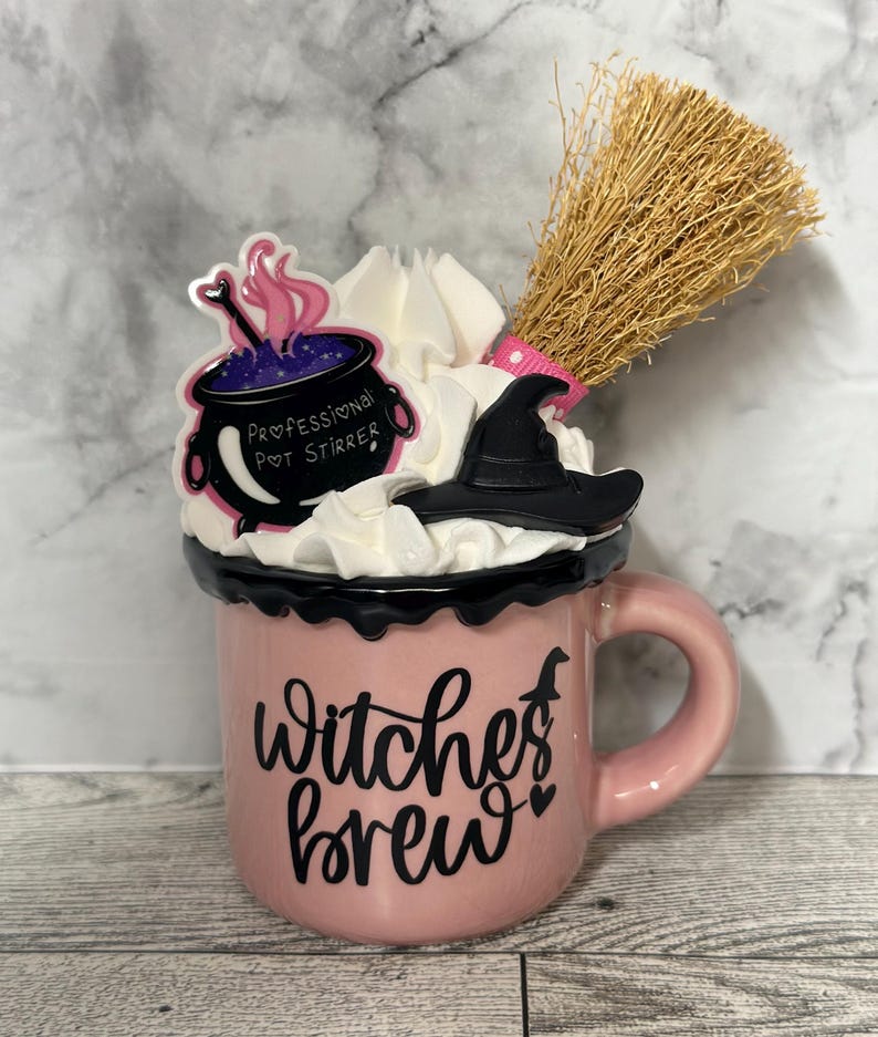 May include: Pink ceramic mug with a black rim and handle. The mug has a black and white graphic that says "Witches Brew" with a small witch silhouette and two hearts. The mug is topped with whipped cream, a black witch's hat, and a small purple cauldron with a pink and white "Professional Pot Stirrer" sticker. A brown broom is sticking out of the top of the mug.