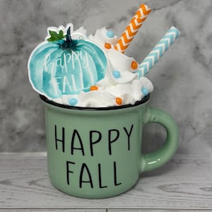 May include: A green ceramic mug with the words "HAPPY FALL" in black. The mug is decorated with whipped cream, a teal pumpkin sticker, and two straws. The pumpkin sticker has the words "happy FALL" in white.