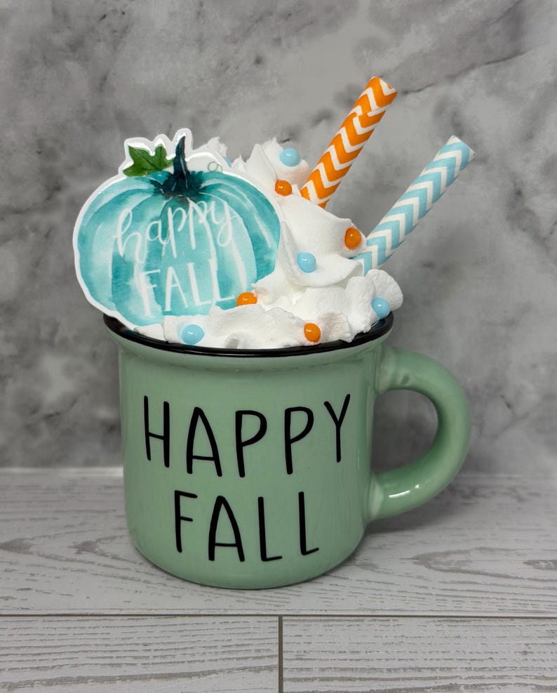 May include: A green ceramic mug with the words "HAPPY FALL" in black. The mug is decorated with whipped cream, a teal pumpkin sticker, and two straws. The pumpkin sticker has the words "happy FALL" in white.