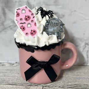 May include: A pink ceramic mug with a black bow, filled with white whipped cream, a pink and white coffin-shaped cookie, a black spider, and a gray tombstone with the word "RIP" on it.