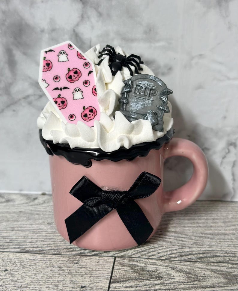 May include: A pink ceramic mug with a black bow, filled with white whipped cream, a pink and white coffin-shaped cookie, a black spider, and a gray tombstone with the word "RIP" on it.