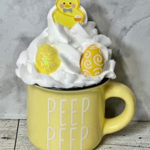 Op de afbeelding: Gele keramische mok met de tekst "PEEP PEEP" aan de voorkant. De mok is gevuld met witte slagroom en bedekt met een geel kuikenbeeldje en twee gele paaseieren.