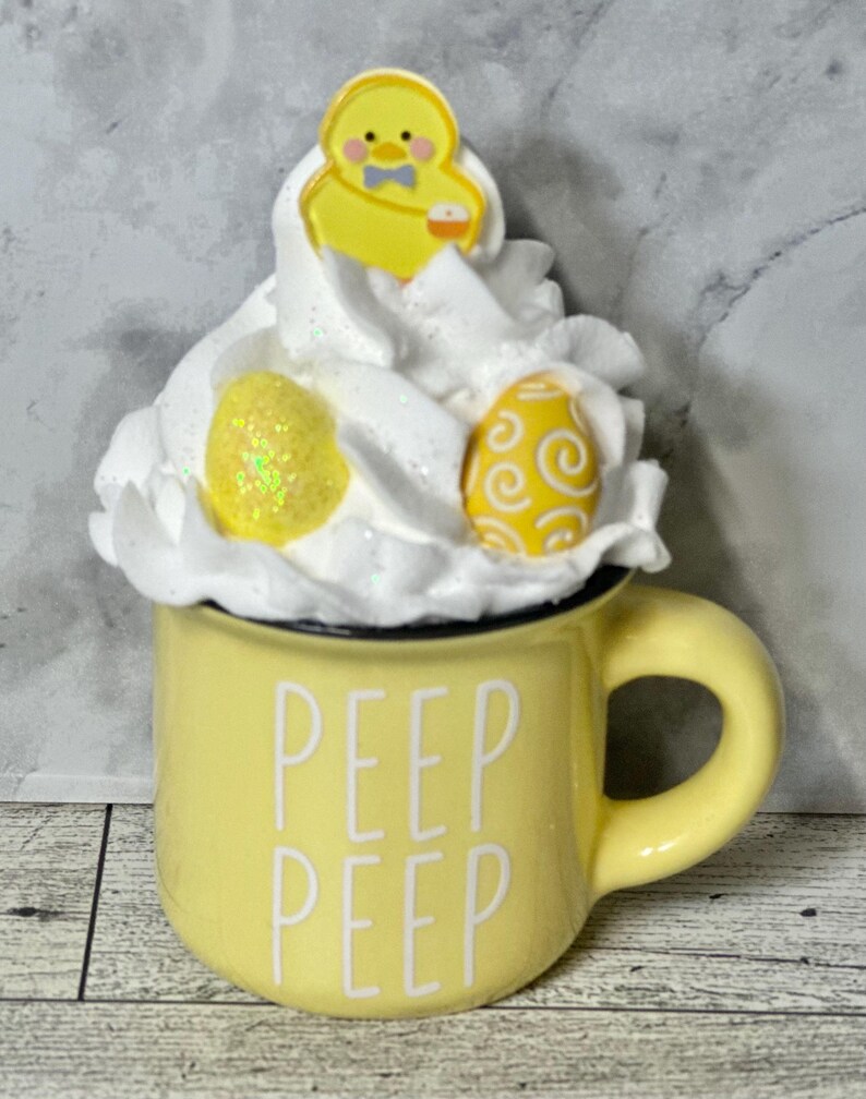 Op de afbeelding: Gele keramische mok met de tekst "PEEP PEEP" aan de voorkant. De mok is gevuld met witte slagroom en bedekt met een geel kuikenbeeldje en twee gele paaseieren.