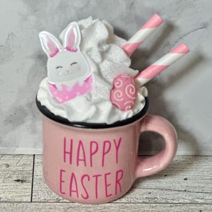 Op de afbeelding: Roze keramische mok met witte slagroom, een roze en wit konijn en een roze en wit swirl snoepje. De mok heeft zwarte letters met de tekst "Happy Easter".