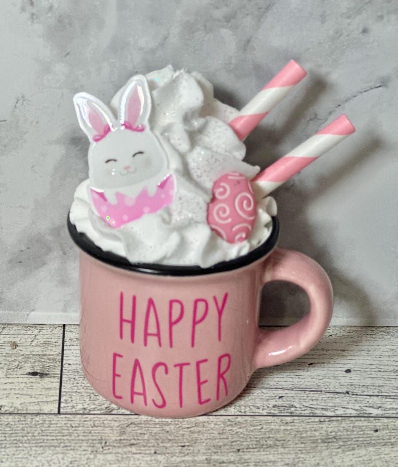 Op de afbeelding: Roze keramische mok met witte slagroom, een roze en wit konijn en een roze en wit swirl snoepje. De mok heeft zwarte letters met de tekst "Happy Easter".