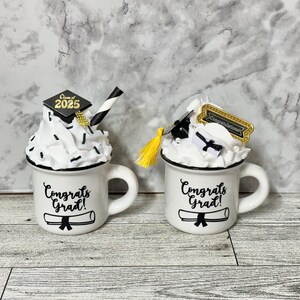 May include: Two white ceramic mugs with black handles. Each mug has a black and white graduation cap and a black and white diploma on it. The text "Congrats Grad!" is printed on each mug.