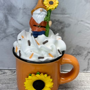 May include: An orange ceramic mug with a black rim, adorned with a yellow sunflower. The mug is topped with white whipped cream and sprinkles, and features a gnome holding a sunflower. A whimsical, decorative item.
