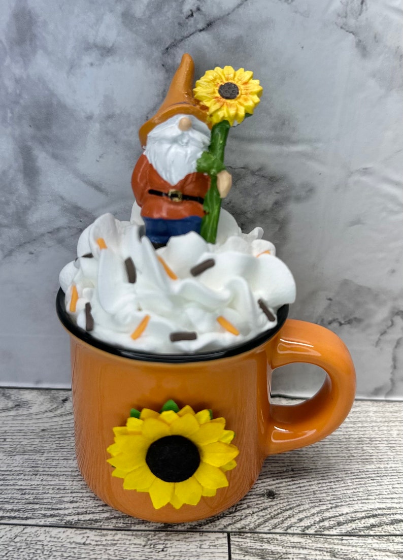 May include: An orange ceramic mug with a black rim, adorned with a yellow sunflower. The mug is topped with white whipped cream and sprinkles, and features a gnome holding a sunflower. A whimsical, decorative item.