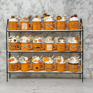 May include: A three-tiered black metal shelf displays orange mugs with whipped cream toppings and decorative accents. Each mug has a different fall-themed message, such as "Gobble Gobble" and "Pumpkin Spice".