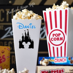 May include: Two white popcorn containers with red and white stripes. One container has a black silhouette of a castle and Mickey Mouse with the name "Daniel" in blue. The other container has the words "POP CORN" in red.