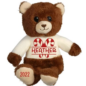 May include: Brown teddy bear wearing a white shirt with a red candy cane design and the name "HEATHER" printed on it. The bear's foot has the year "2022" printed on it.