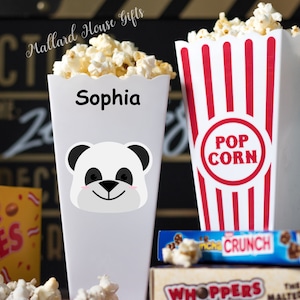 Może przedstawiać: Dwa białe pojemniki na popcorn w czerwono-białe paski. Jeden pojemnik ma twarz pandy i napis "Sophia" nadrukowany na nim. Drugi pojemnik ma napis "POP CORN" nadrukowany na nim.