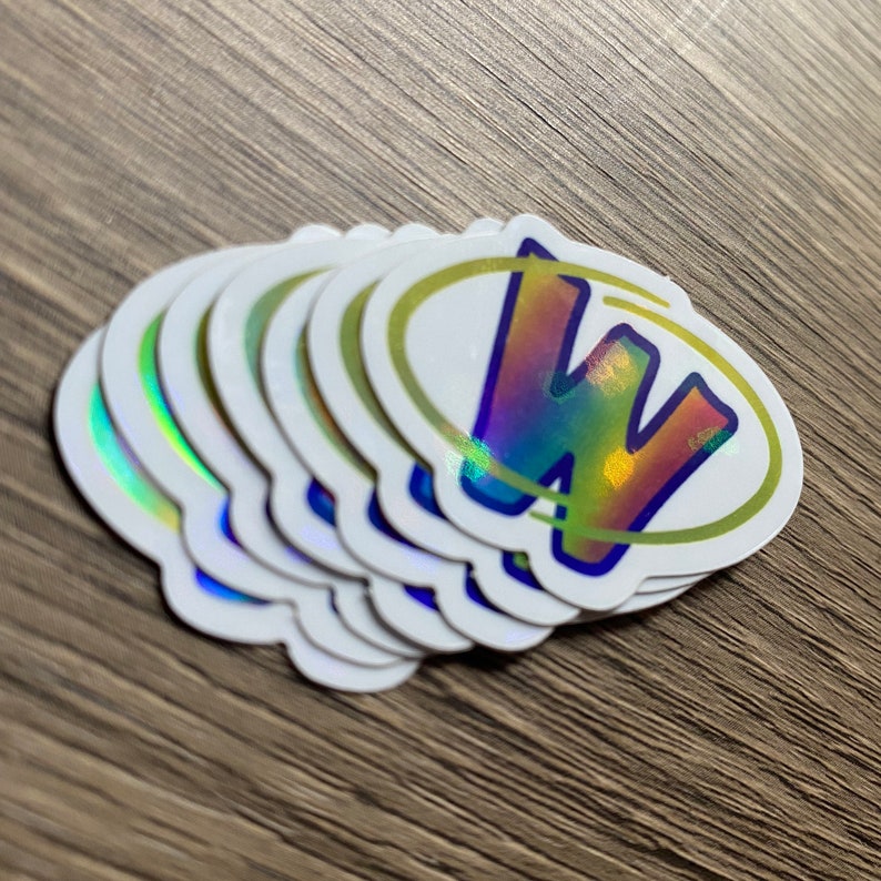 K&ouml;nnte beinhalten: Ein Stapel von acht schimmernden wei&szlig;en ovalen Aufklebern mit gelbem Rand und einem holografischen blauen und regenbogenfarbenen Buchstaben "W" darin.