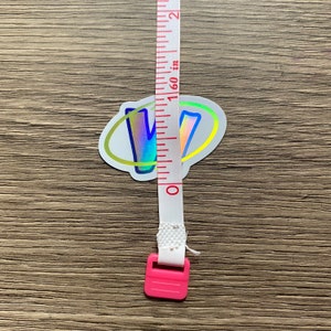 K&ouml;nnte beinhalten: Ein holografischer Aufkleber mit dem Buchstaben "V" in einem Regenbogen-Farbschema, umgeben von einem gelben Kreis. Der Aufkleber wird mit einem wei&szlig;en Ma&szlig;band mit rosa Endkappen gemessen.