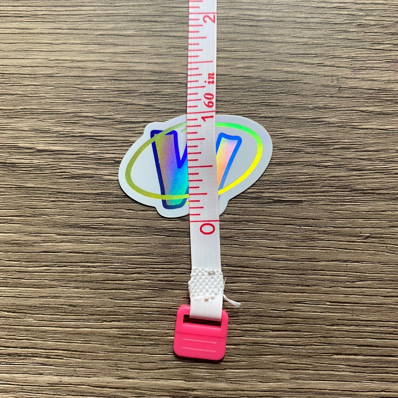 K&ouml;nnte beinhalten: Ein holografischer Aufkleber mit dem Buchstaben "V" in einem Regenbogen-Farbschema, umgeben von einem gelben Kreis. Der Aufkleber wird mit einem wei&szlig;en Ma&szlig;band mit rosa Endkappen gemessen.