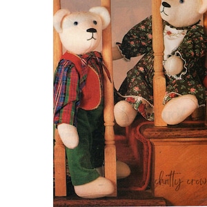 Può includere: Due orsacchiotti bianchi vestiti con abiti vintage. L'orso a sinistra indossa una camicia a quadri, un gilet rosso e pantaloni verdi. L'orso a destra indossa un vestito floreale. Gli orsi sono in piedi su una scala di legno con il testo "chatty crow" visibile in basso.