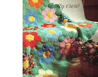 May include: A crocheted blanket with a floral design in shades of green, blue, yellow, red, and pink. The blanket is made up of individual flower motifs, each with a different color combination. The blanket is draped over a couch and a potted plant is in the foreground.