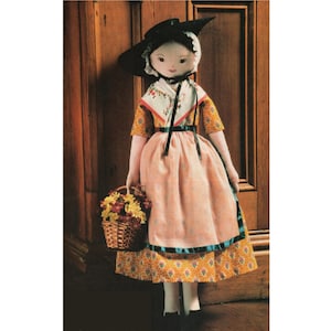 May include: A handmade doll wearing a pink dress with a floral print, a black hat, and a white lace collar. The doll is holding a basket of yellow flowers.