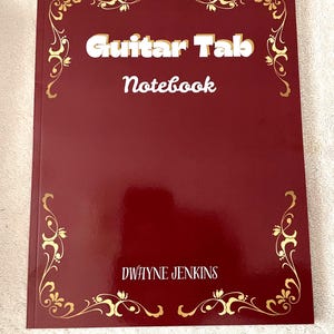 May include: A burgundy Guitar Tab Notebook with gold decorative flourishes. The words "Guitar Tab Notebook" are in white, with the name "Dwayne Jenkins" at the bottom. The notebook has a glossy finish.