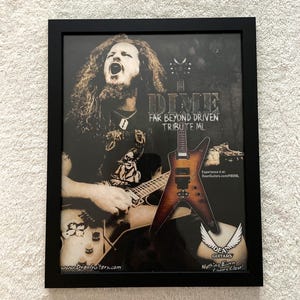 May include: A black framed poster featuring a photograph of Dimebag Darrell playing a Dean guitar. The text reads DIME FAR BEYOND DRIVEN TRIBUTE ML, Experience it at: DeanGuitars.com/FBDML, Dean Guitars, Nothing Even Comes Close!, and www.DeanGuitars.com.