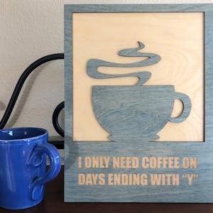 Puede incluir: Un letrero de madera con fondo gris y marco de madera clara. El letrero presenta una silueta de una taza de café con vapor saliendo de ella. El texto en el letrero dice "Solo necesito café en los días que terminan en "Y"".