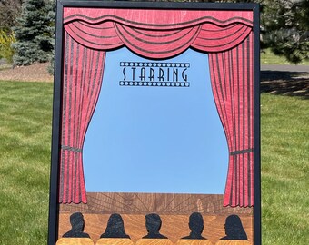 May include: A wooden theater-themed sign with a black frame. The sign features red curtains, a blue backdrop, and the word "STARRING" at the top. Below the stage are silhouettes of people in the audience. The sign is displayed on a black easel.