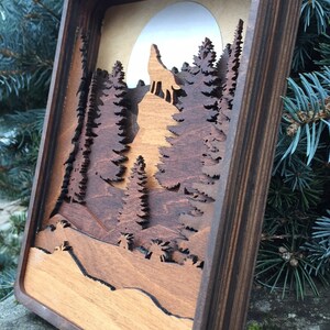 May include: A wooden wall art piece with a layered design of a wolf howling at the moon in a forest setting. The piece is framed in dark wood and has a natural wood finish.