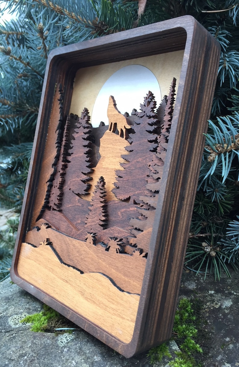 May include: A wooden wall art piece with a layered design of a wolf howling at the moon in a forest setting. The piece is framed in dark wood and has a natural wood finish.